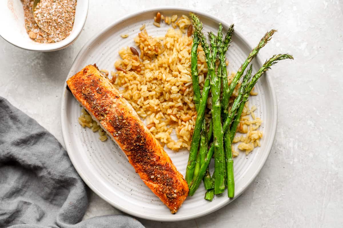 Close-up view of seasoned salmon, rice pilaf, and asparagus on a white plate with a gold fork.