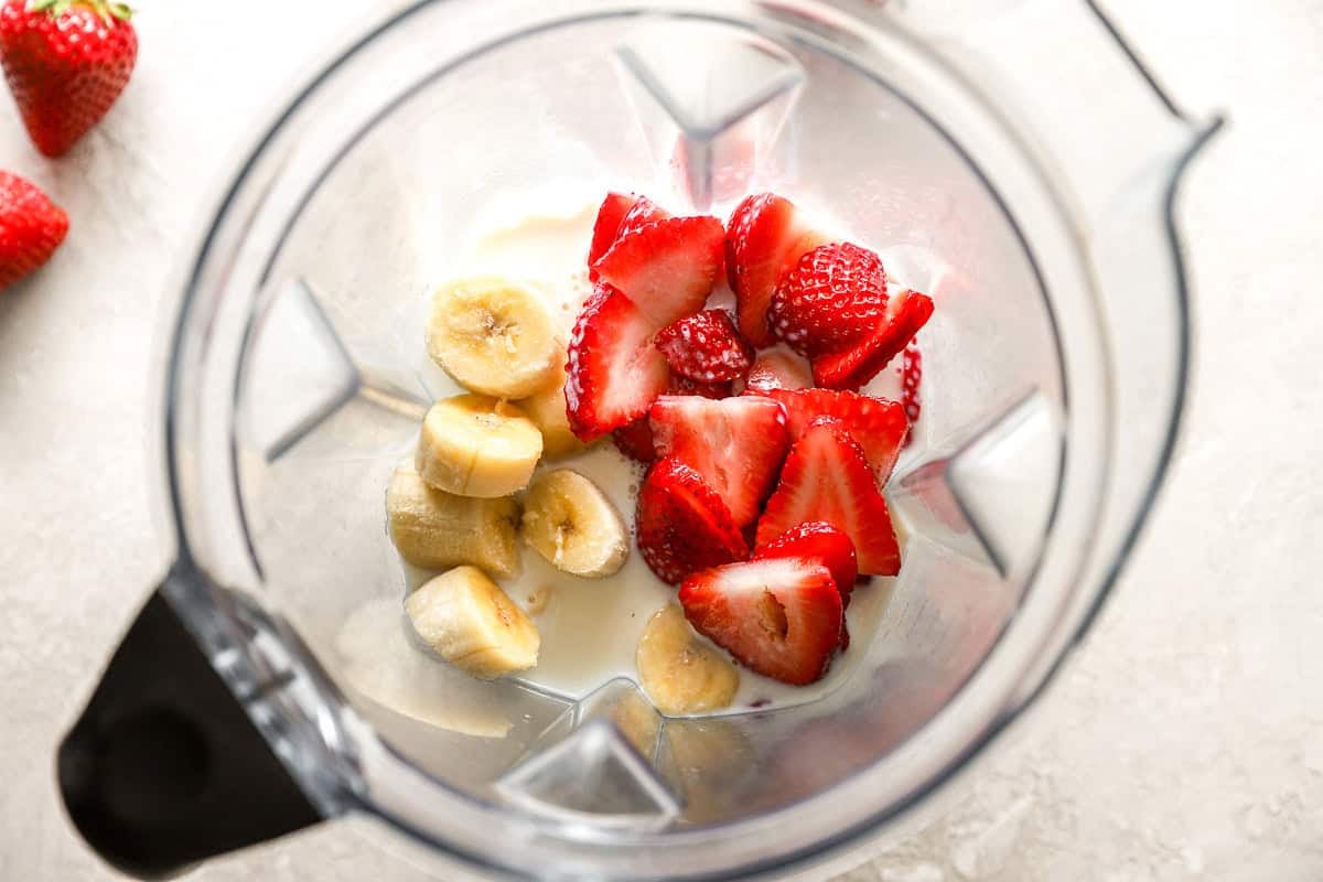 Overhead view of a blender containing sliced bananas, strawberries, and milk, ready to be blended into a smoothie.