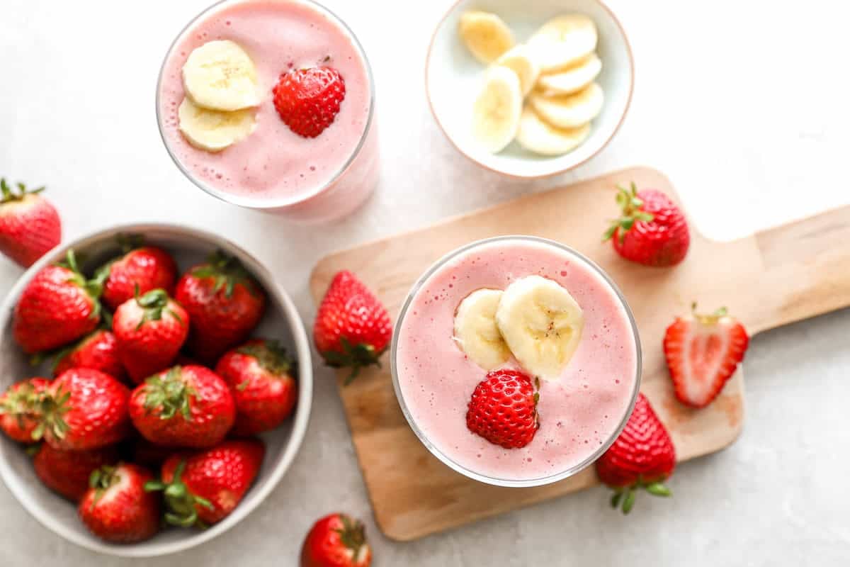 Overhead view of two glasses of strawberry banana smoothie topped with banana slices and strawberries, surrounded by fresh fruit on a light surface.