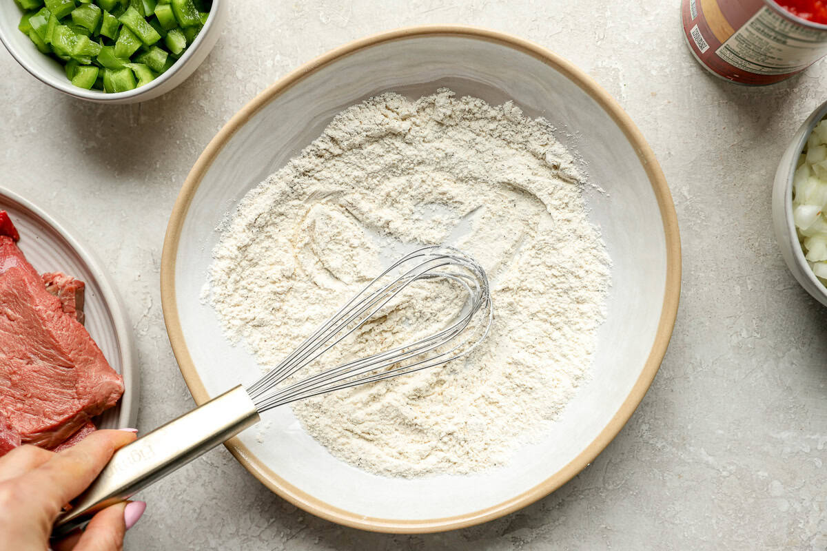 Bowl with whisked flour, onion powder, garlic powder, salt, and pepper mixture for dredging steak. Ingredients like green pepper, steak, and onion surround the bowl.