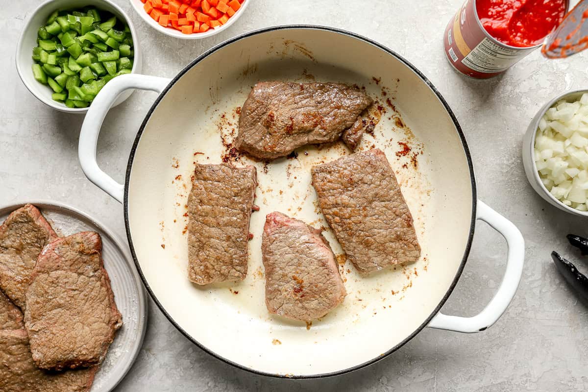 Round steak slices browning in a large white skillet with bowls of chopped vegetables and crushed tomatoes nearby.