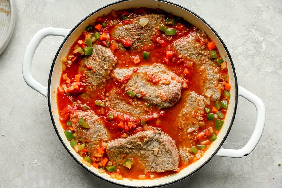 Steak pieces simmering in a rich tomato and vegetable sauce inside a large white skillet.