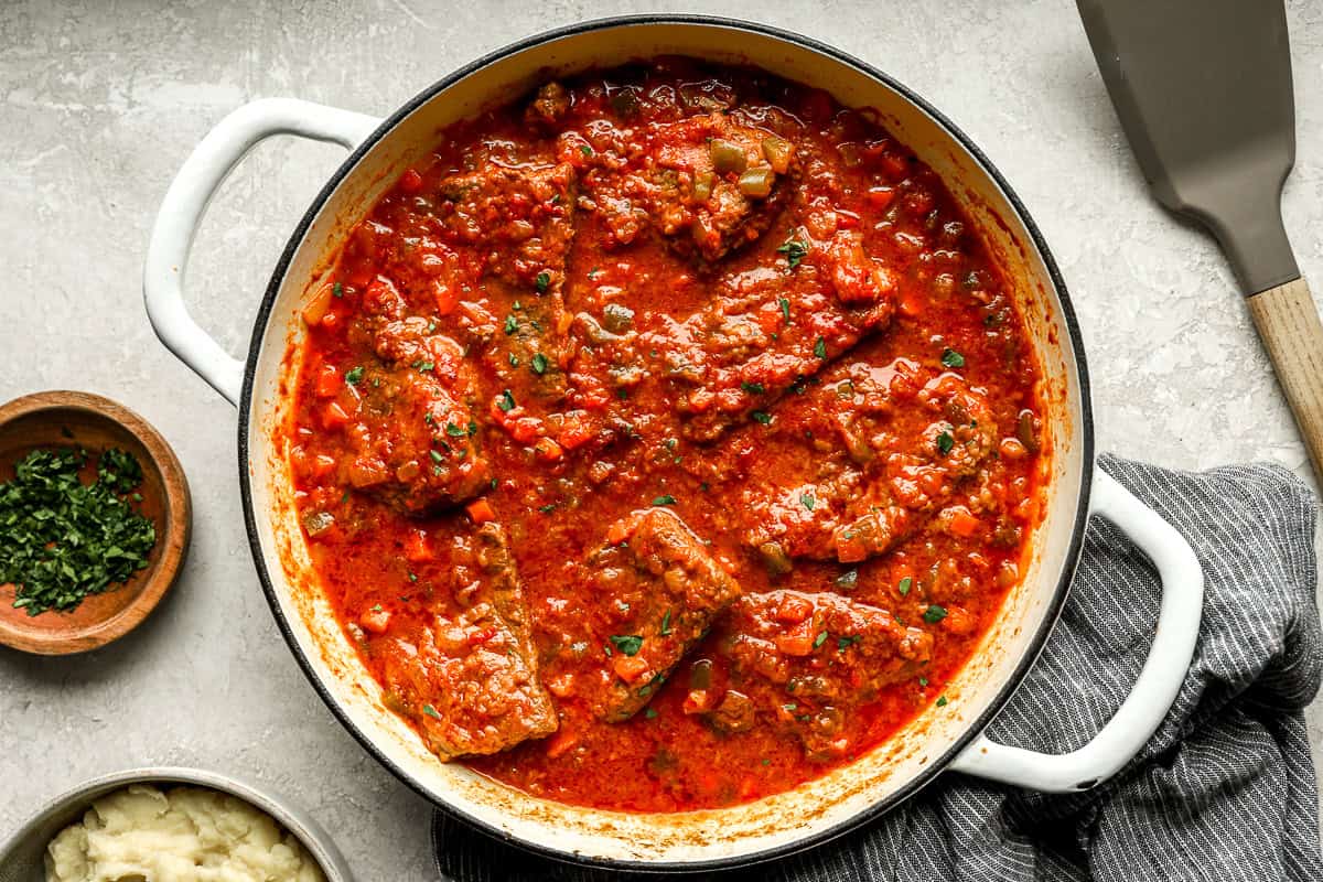Swiss steak fully cooked in tomato sauce, garnished with parsley, inside a white Dutch oven with a dish towel and chopped herbs nearby.