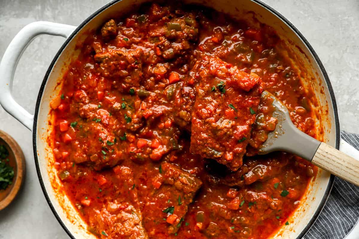 Close-up of a spatula lifting a tender piece of Swiss steak from a skillet filled with chunky tomato sauce and vegetables.