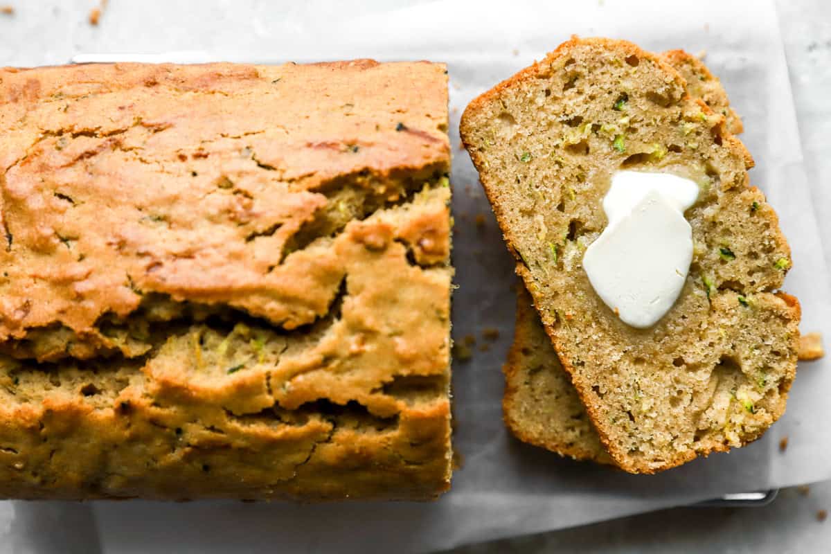 Close-up of a slice of zucchini bread topped with a pat of melting butter, next to the full loaf.