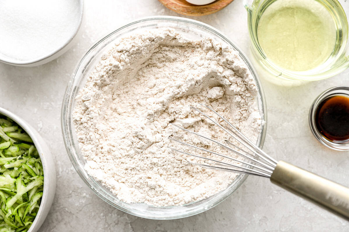Overhead view of dry ingredients for zucchini bread in a glass bowl with a whisk, surrounded by bowls of sugar, oil, vanilla, and grated zucchini.