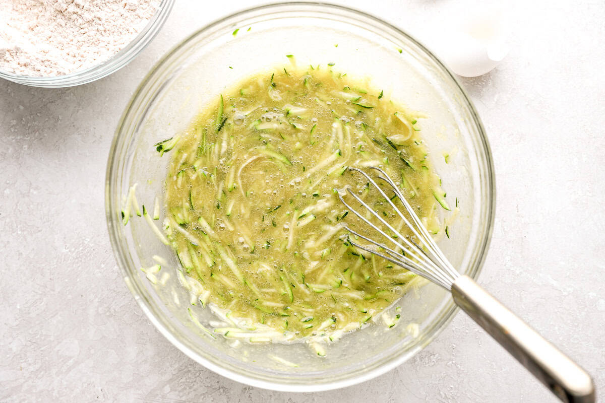 Wet ingredients mixed with shredded zucchini in a glass bowl, with a whisk resting in the mixture.