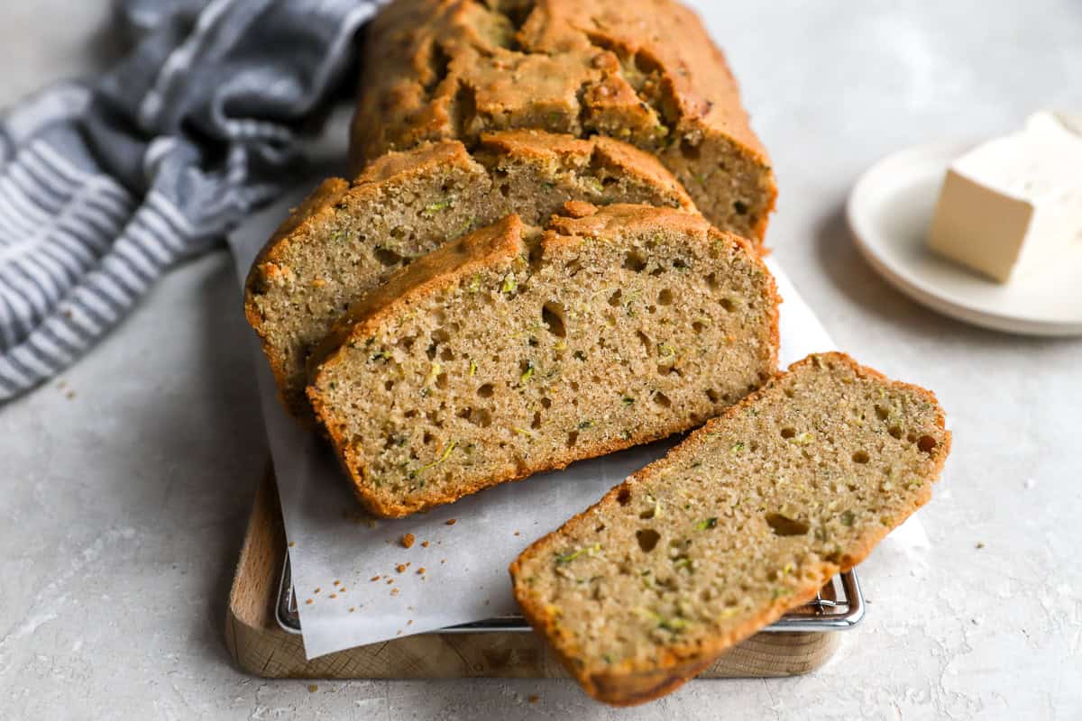 Sliced zucchini bread displayed on a cutting board with parchment paper, next to a striped napkin and a plate of butter.