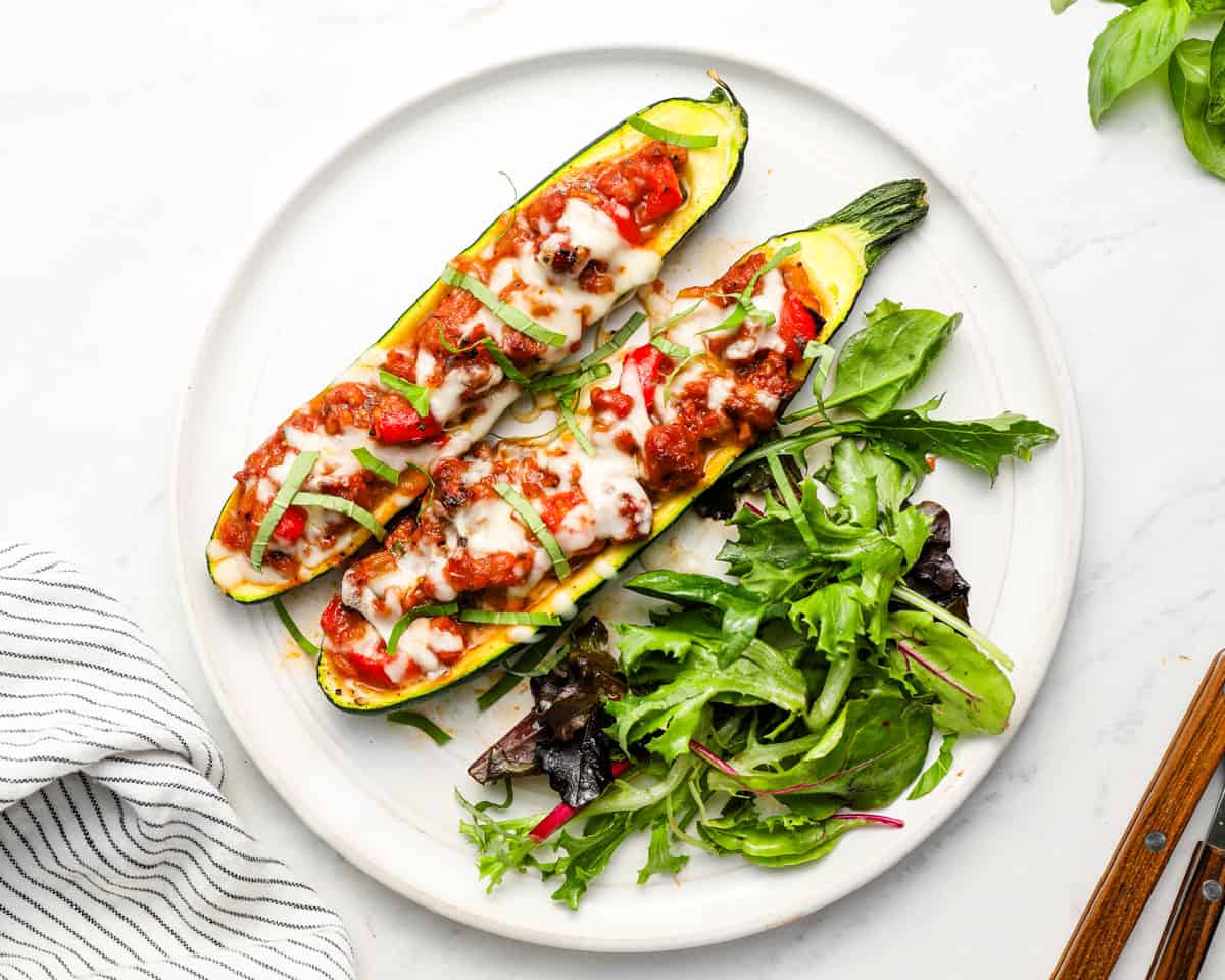 Two sausage stuffed zucchini boats topped with melted mozzarella and fresh basil, served with a side of mixed greens on a white plate.