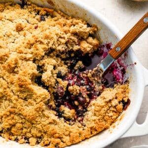 Close-up of a baked blueberry crisp in a white oval dish with a spoon scooping out the juicy berry filling and golden crumble topping.