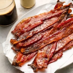 Plate of crispy candied bacon strips on parchment paper served with coffee, milk, and fresh strawberries.