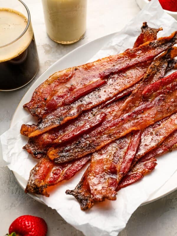 Plate of crispy candied bacon strips on parchment paper served with coffee, milk, and fresh strawberries.