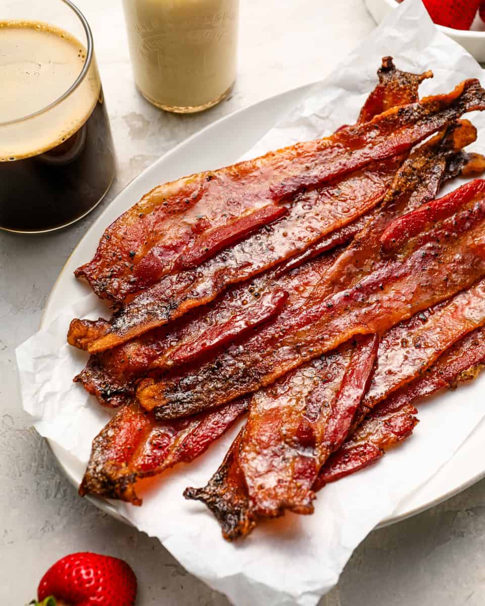Plate of crispy candied bacon strips on parchment paper served with coffee, milk, and fresh strawberries.