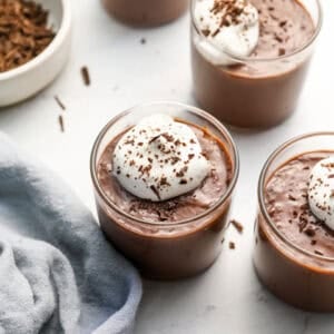 Finished chocolate pudding in small glass cups topped with whipped cream and chocolate shavings, placed on a light surface with a bowl of shaved chocolate and a linen napkin nearby.