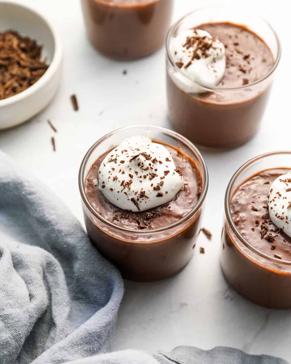 Finished chocolate pudding in small glass cups topped with whipped cream and chocolate shavings, placed on a light surface with a bowl of shaved chocolate and a linen napkin nearby.