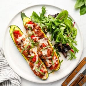 Two stuffed zucchini boats topped with melted cheese and fresh basil served with a side salad on a white plate.