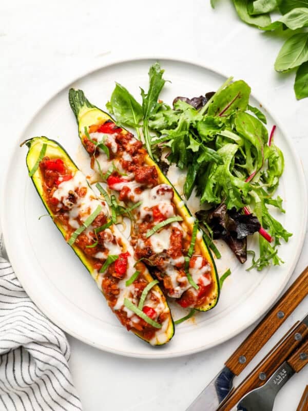 Two stuffed zucchini boats topped with melted cheese and fresh basil served with a side salad on a white plate.