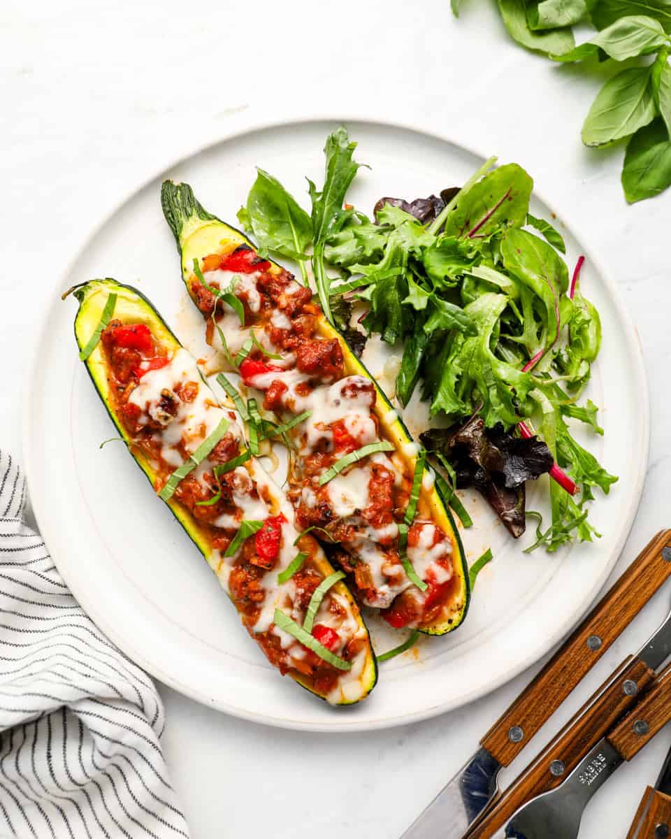 Two stuffed zucchini boats topped with melted cheese and fresh basil served with a side salad on a white plate.