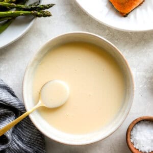 Creamy beurre blanc sauce in a bowl with a gold spoon, next to roasted asparagus and salmon on a white plate.