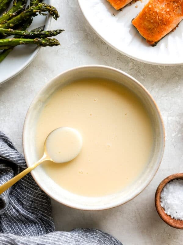 Creamy beurre blanc sauce in a bowl with a gold spoon, next to roasted asparagus and salmon on a white plate.