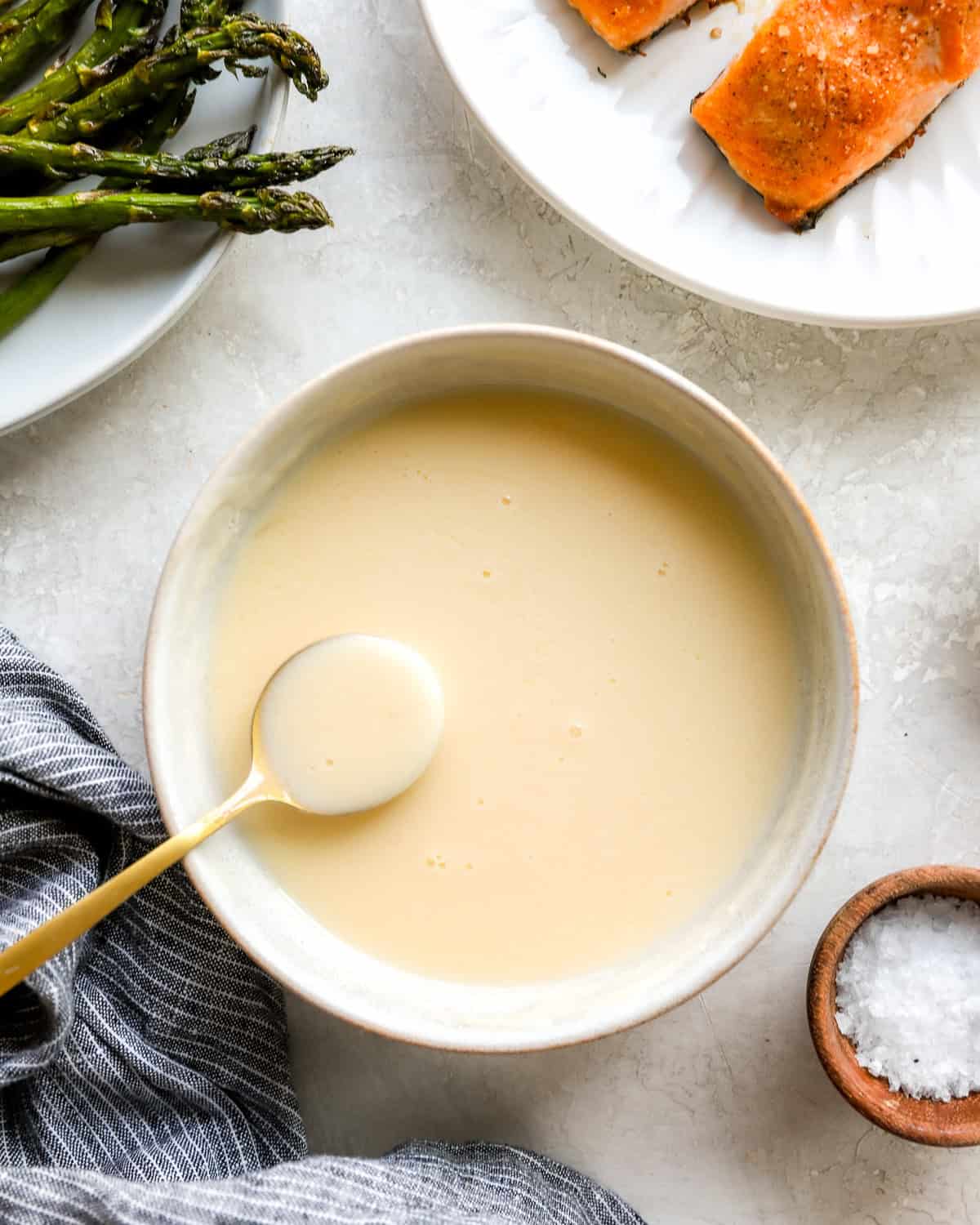 Creamy beurre blanc sauce in a bowl with a gold spoon, next to roasted asparagus and salmon on a white plate.