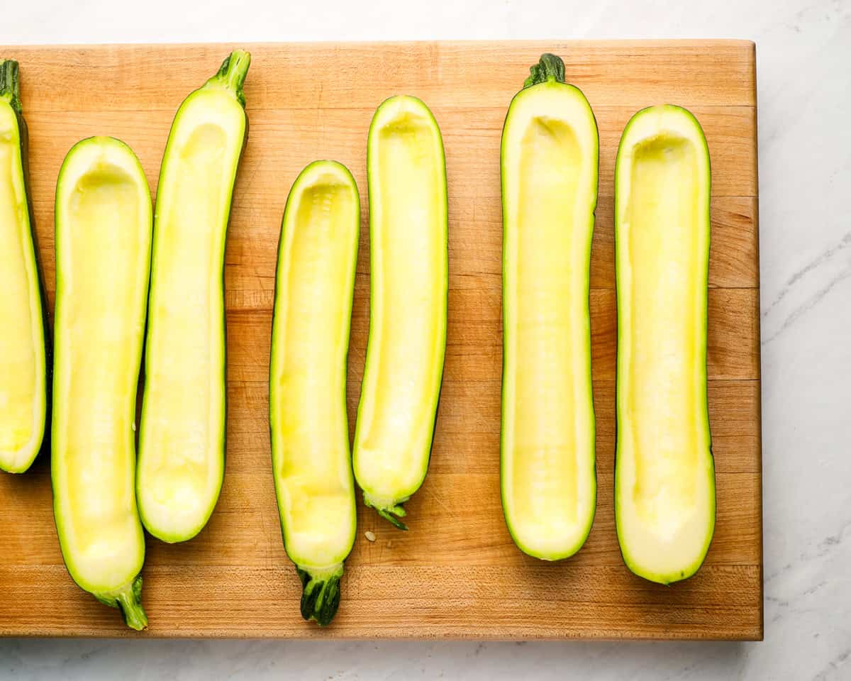 Eight zucchini halves, hollowed out and arranged on a wooden cutting board, ready to be stuffed.