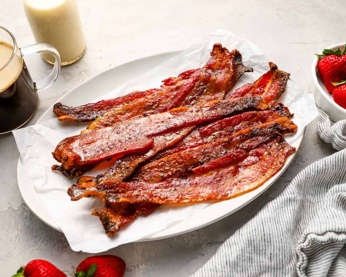 Plated candied bacon on white parchment with coffee, milk, strawberries, and a striped towel for serving.