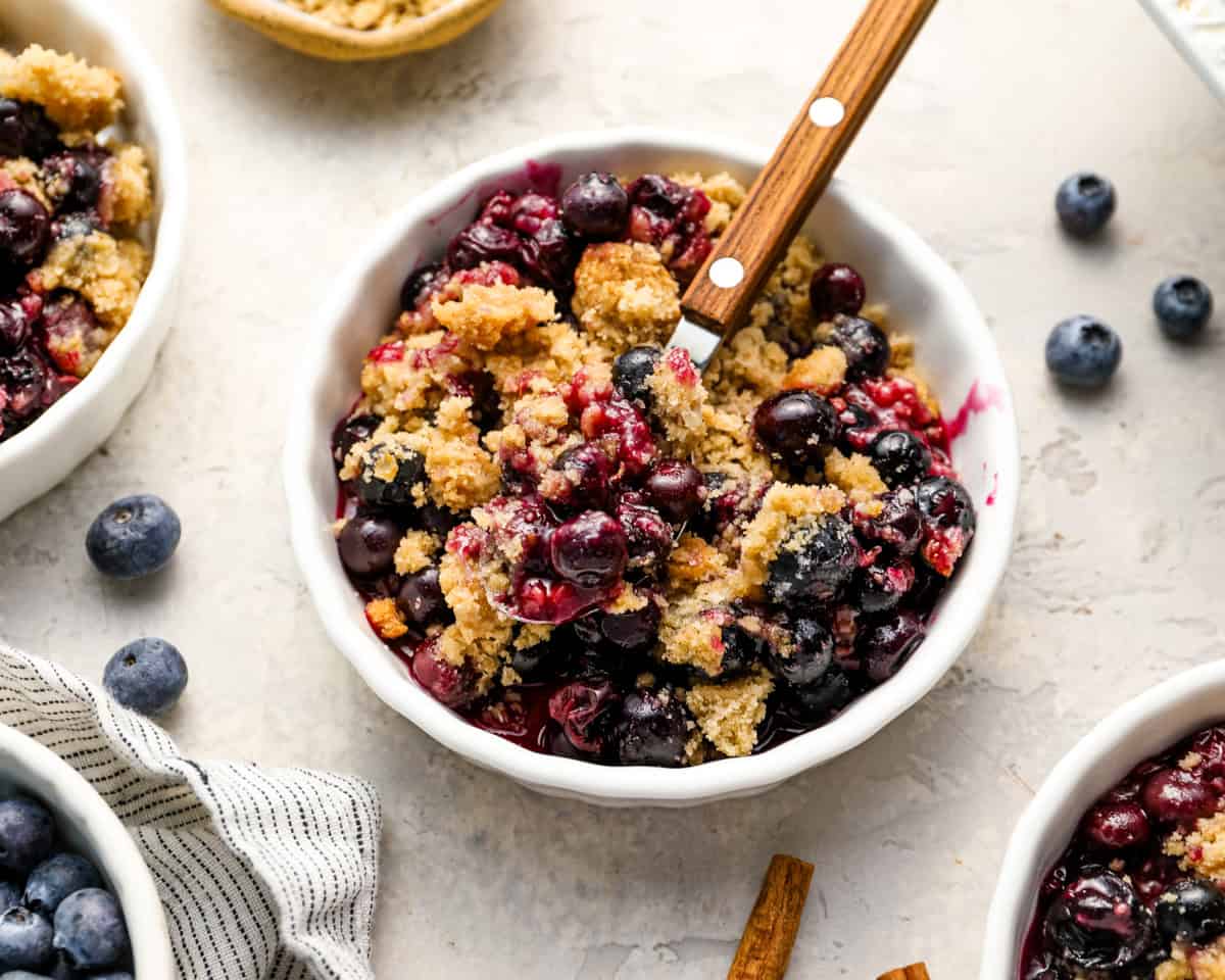 Serving of blueberry crisp in a small white bowl with a wooden-handled spoon and whole blueberries scattered around.