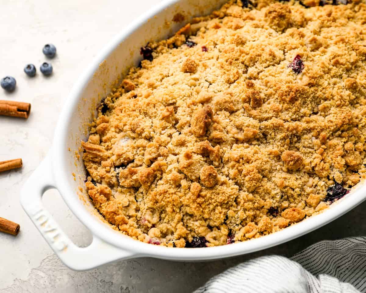 Side view of a golden, bubbling blueberry crisp in a white baking dish with cinnamon sticks and blueberries around.