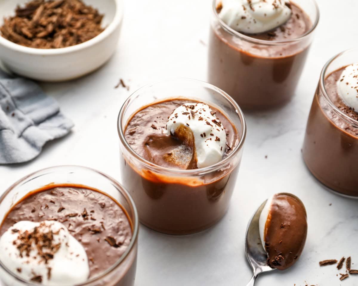 Chocolate pudding cup with a spoonful scooped out, showing the creamy texture. Whipped cream and chocolate shavings are on top, with a spoon resting nearby.