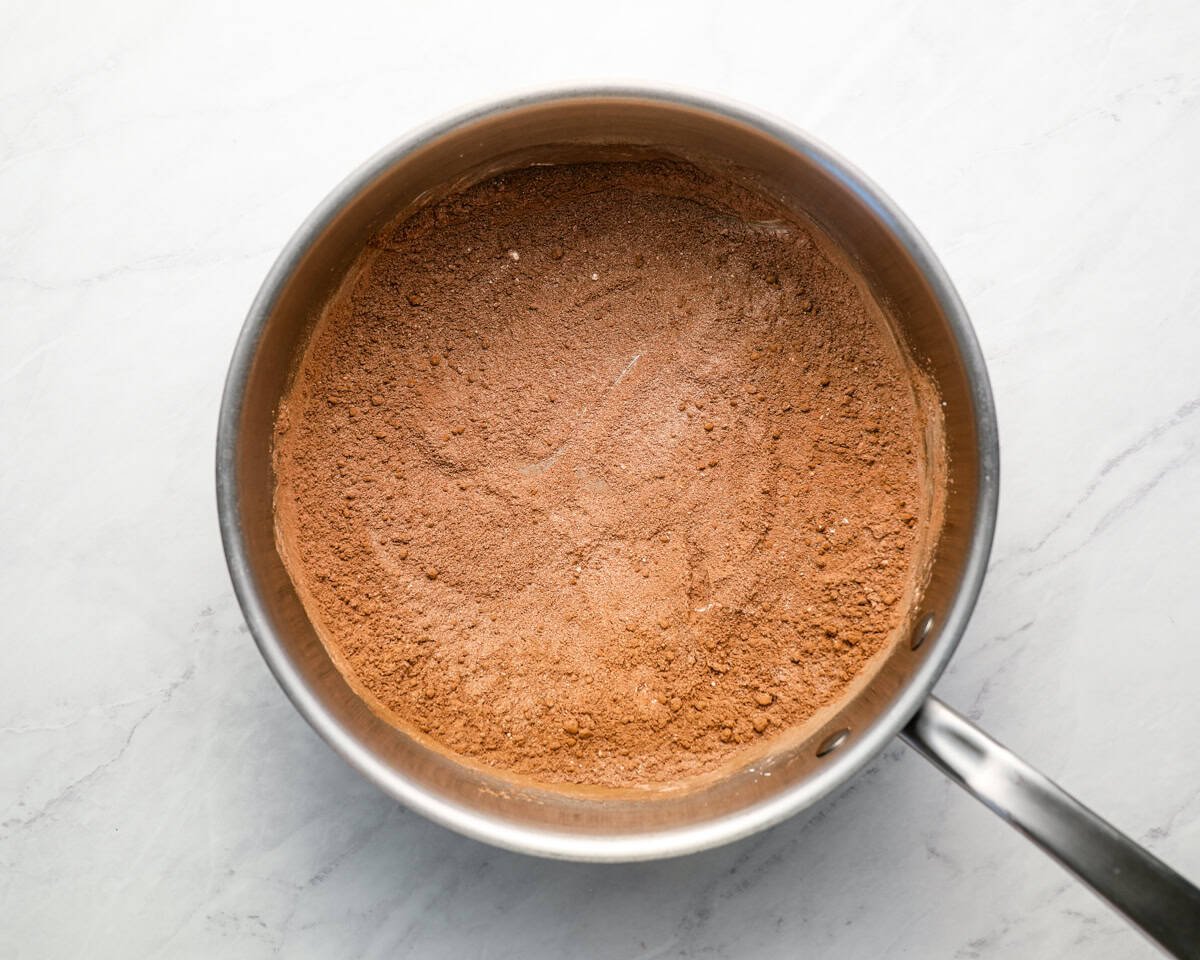 Dry ingredients for chocolate pudding (cocoa powder, sugar, cornstarch, and salt) whisked together in a stainless steel saucepan on a marble surface.