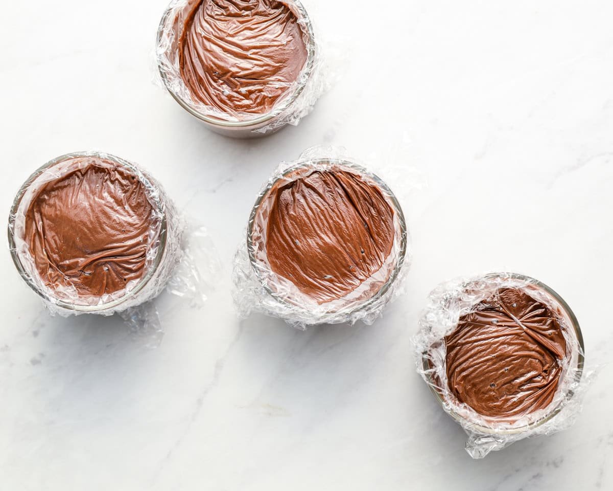 Chocolate pudding poured into small glass bowls with plastic wrap pressed directly onto the surface to prevent a skin from forming.
