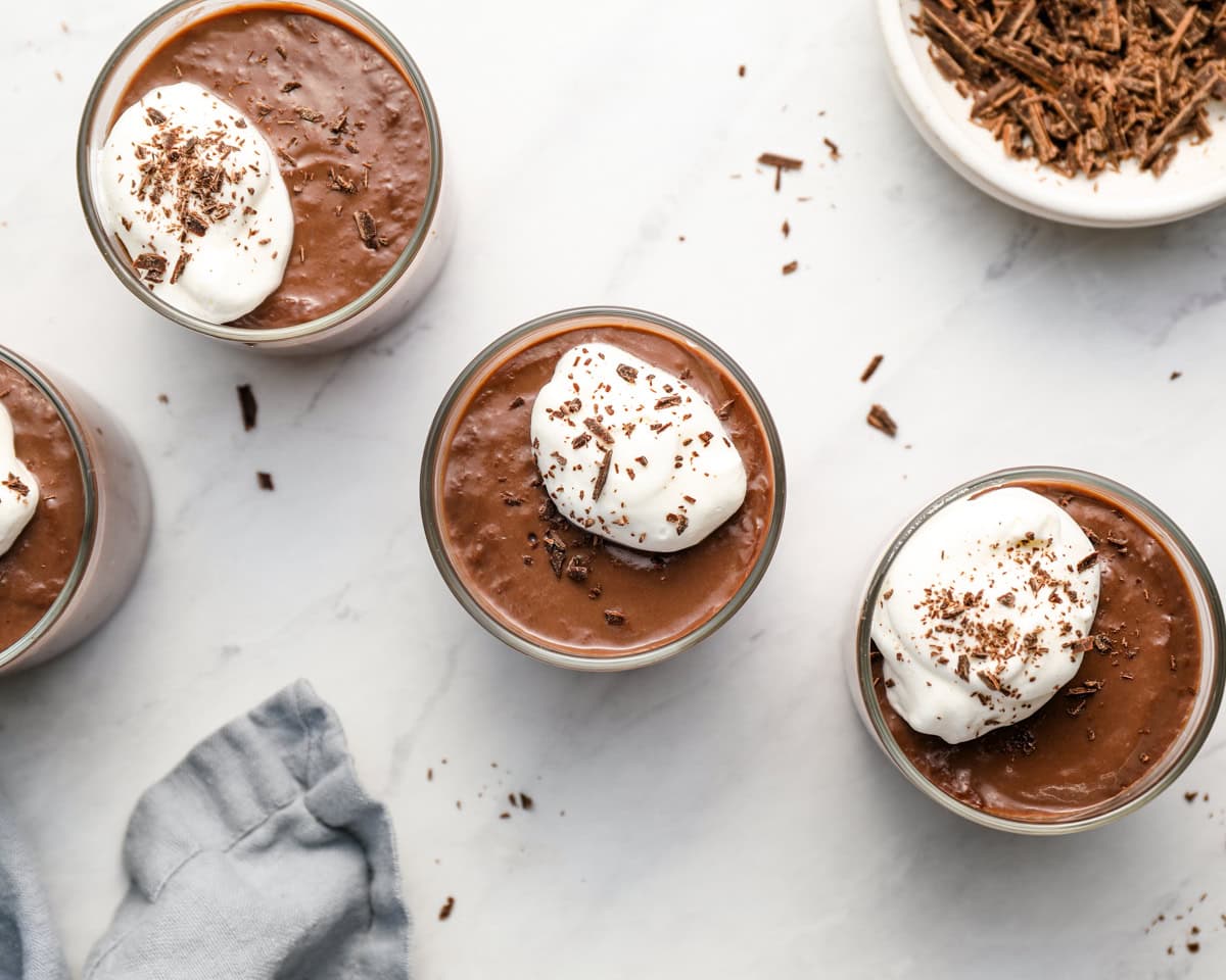 Overhead view of chilled chocolate pudding in serving cups, topped with whipped cream and chocolate shavings.