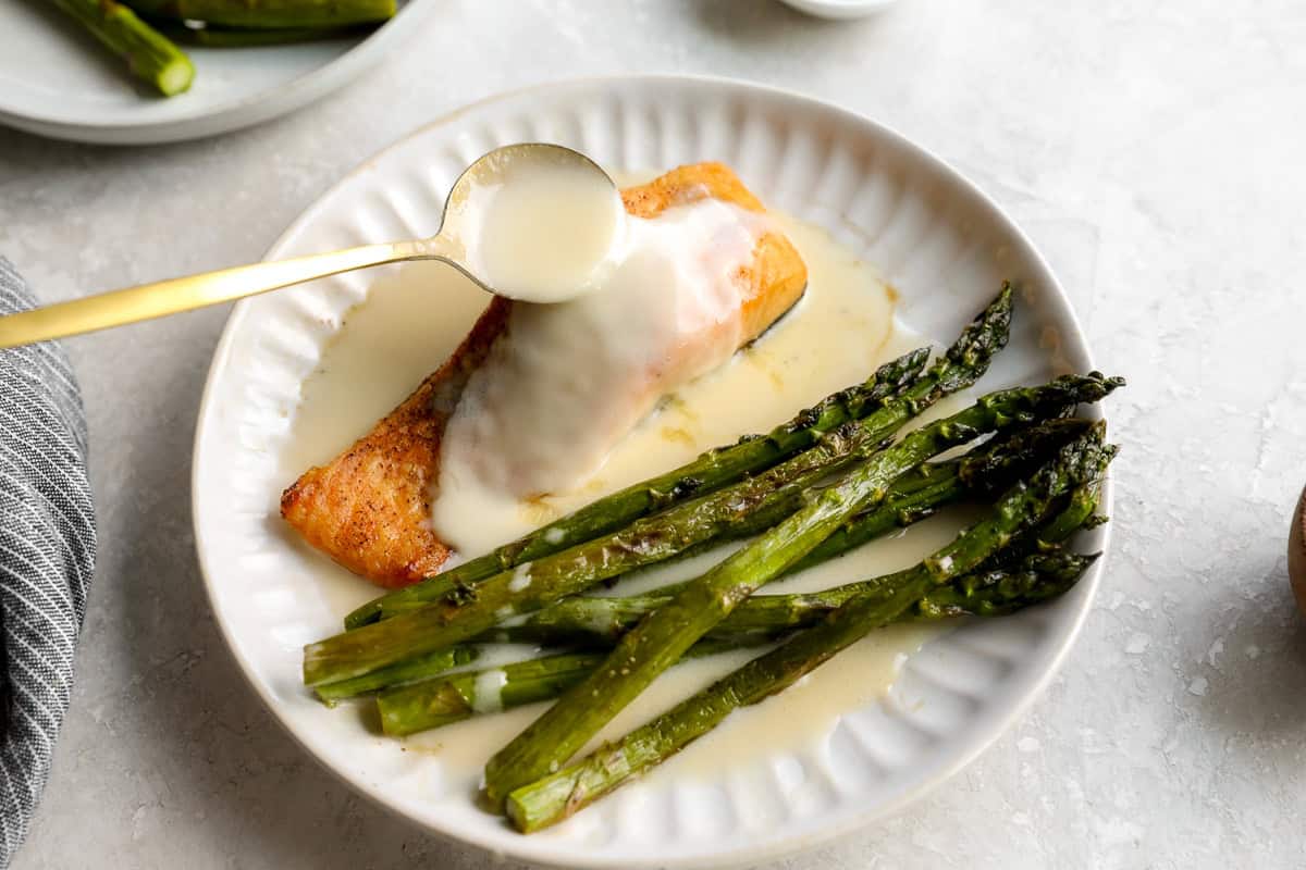 Spoonful of beurre blanc sauce being poured over salmon with roasted asparagus.