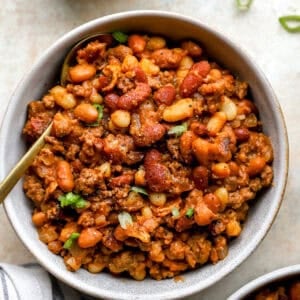 Overhead view of a bowl filled with hearty cowboy beans made with mixed beans, ground beef, and bacon, garnished with chopped green onions and served with a gold spoon.