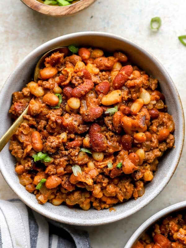 Overhead view of a bowl filled with hearty cowboy beans made with mixed beans, ground beef, and bacon, garnished with chopped green onions and served with a gold spoon.