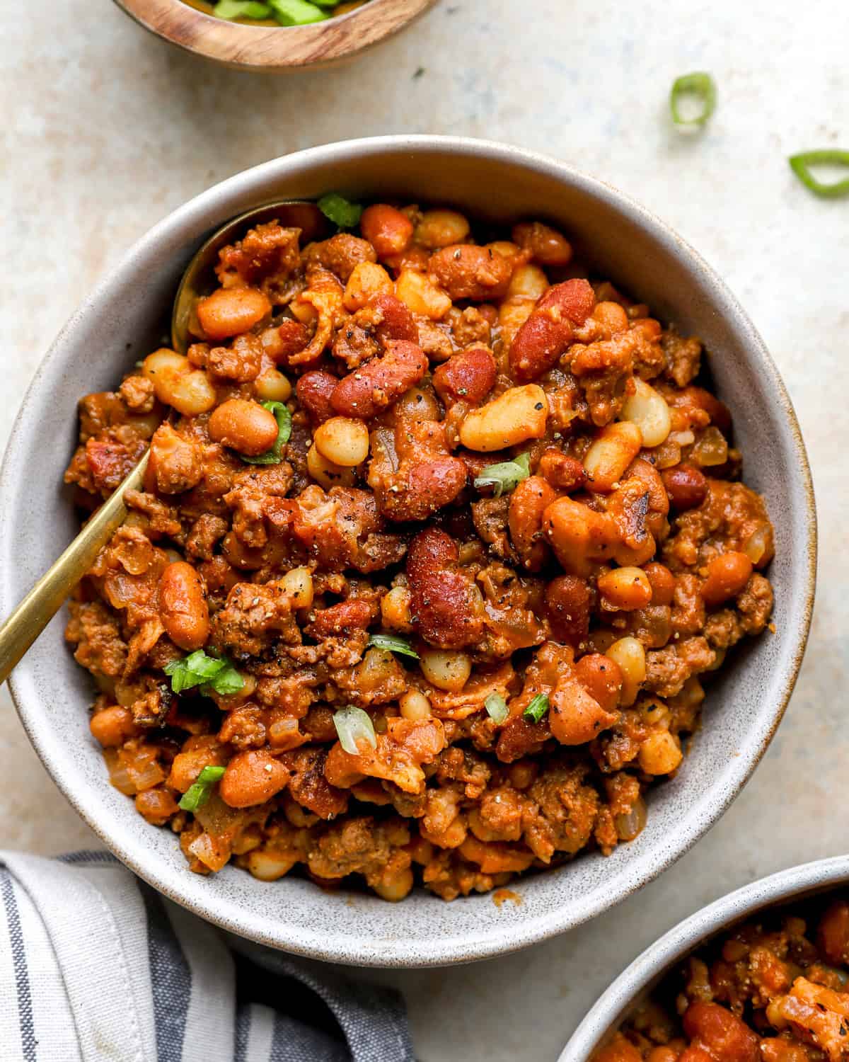 Overhead view of a bowl filled with hearty cowboy beans made with mixed beans, ground beef, and bacon, garnished with chopped green onions and served with a gold spoon.