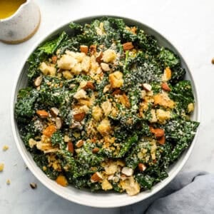 Overhead shot of kale salad in a white bowl topped with homemade breadcrumbs, chopped almonds, and grated parmesan cheese.
