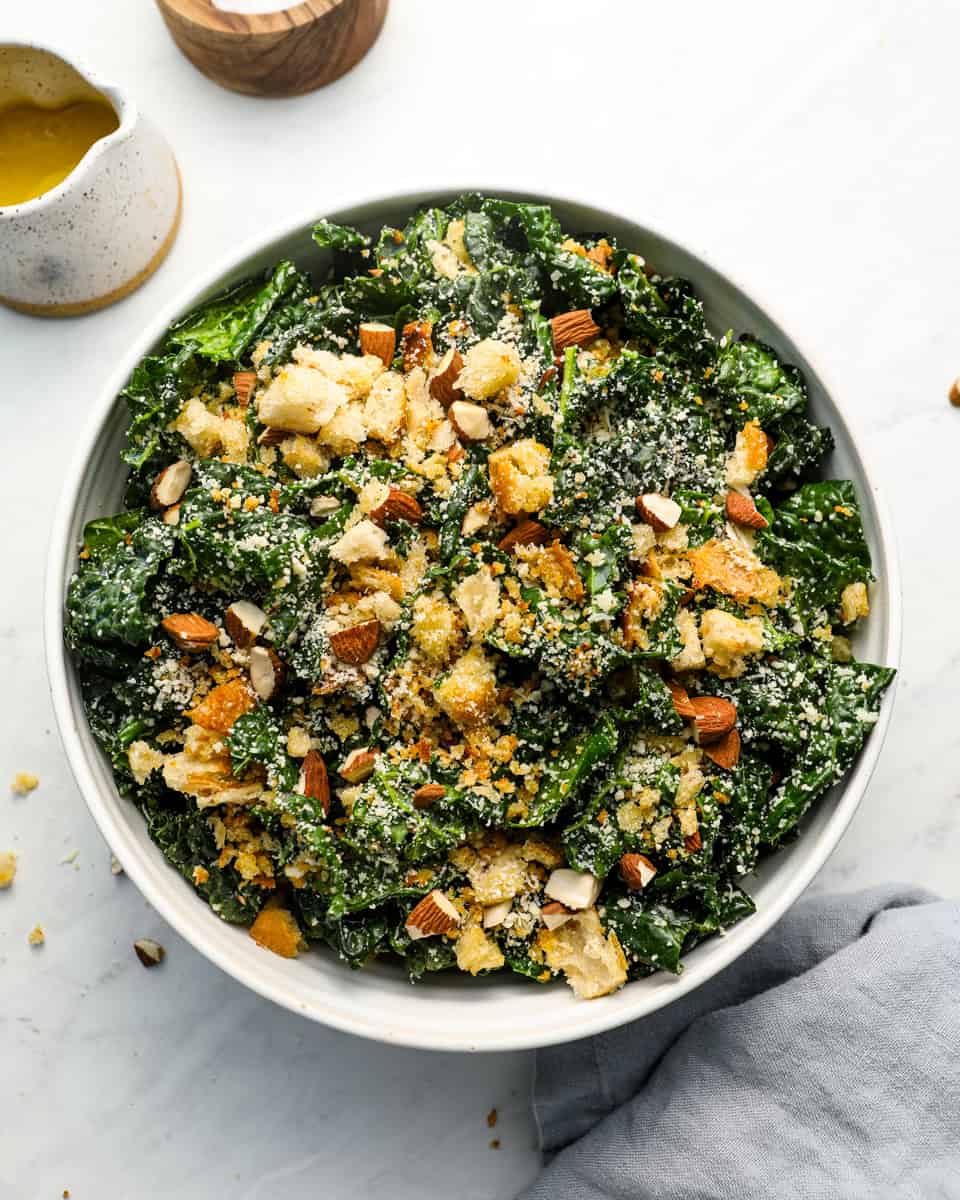 Overhead shot of kale salad in a white bowl topped with homemade breadcrumbs, chopped almonds, and grated parmesan cheese.