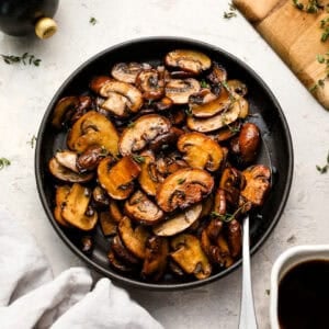 Overhead shot of sautéed mushrooms in a black bowl, garnished with fresh thyme, with a spoon resting inside the bowl.