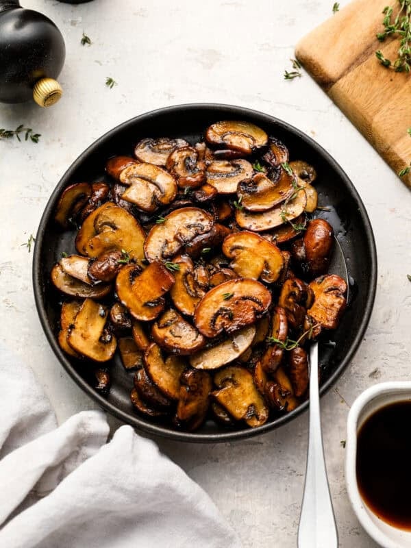 Overhead shot of sautéed mushrooms in a black bowl, garnished with fresh thyme, with a spoon resting inside the bowl.