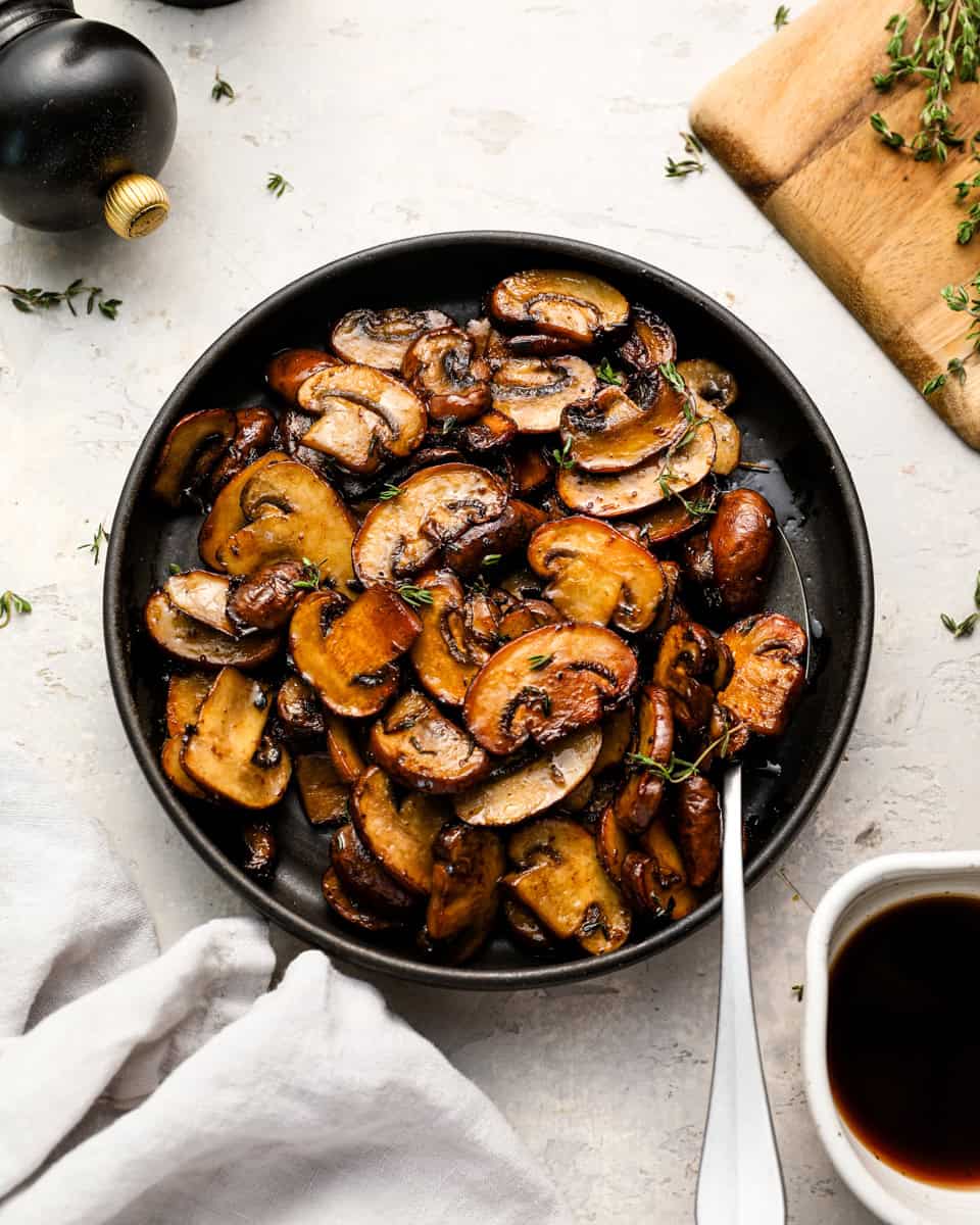 Overhead shot of sautéed mushrooms in a black bowl, garnished with fresh thyme, with a spoon resting inside the bowl.