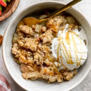 Bowl of apple crumble topped with a scoop of vanilla ice cream and drizzled with caramel sauce, served with a gold spoon.