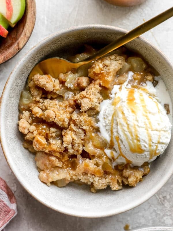 Bowl of apple crumble topped with a scoop of vanilla ice cream and drizzled with caramel sauce, served with a gold spoon.