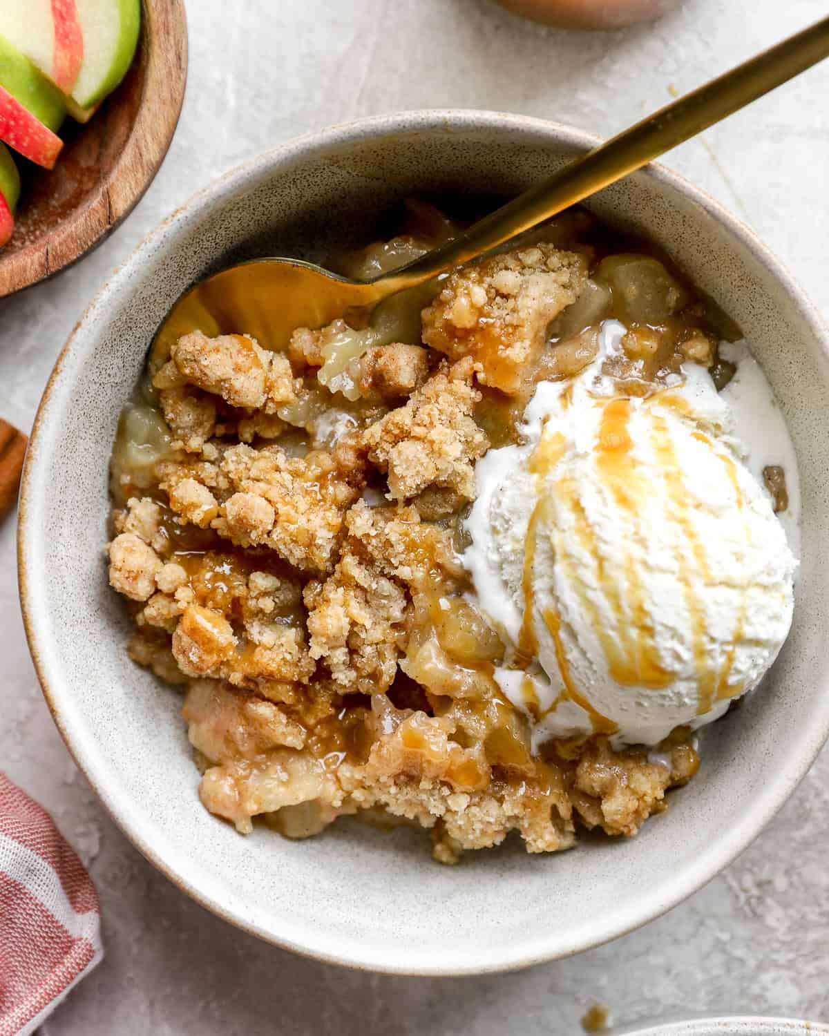 Bowl of apple crumble topped with a scoop of vanilla ice cream and drizzled with caramel sauce, served with a gold spoon.