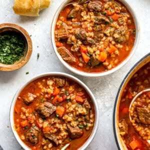 Two bowls of hearty beef barley soup filled with tender beef, carrots, celery, and barley in a rich broth, served with crusty bread and fresh parsley.