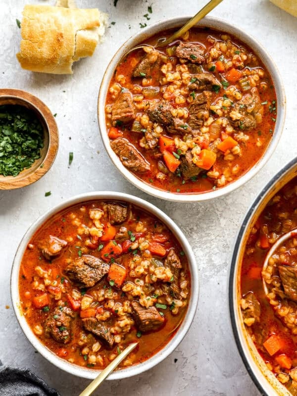 Two bowls of hearty beef barley soup filled with tender beef, carrots, celery, and barley in a rich broth, served with crusty bread and fresh parsley.