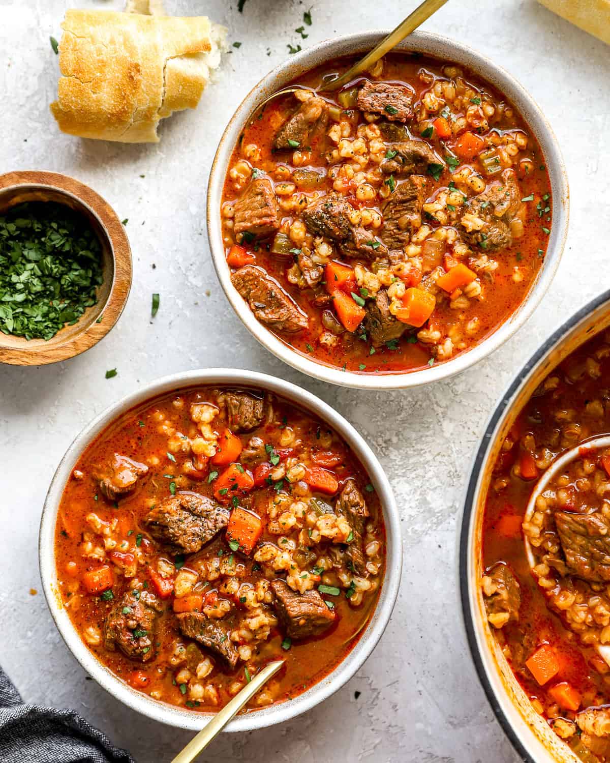 Two bowls of hearty beef barley soup filled with tender beef, carrots, celery, and barley in a rich broth, served with crusty bread and fresh parsley.