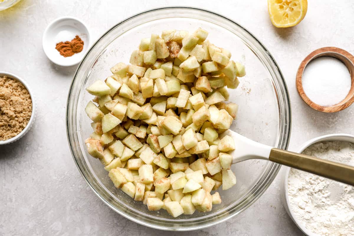Large glass mixing bowl filled with chopped Granny Smith apples coated with cinnamon and sugar, surrounded by small bowls of ingredients.