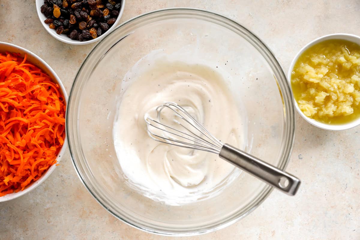 Mixing bowl with creamy dressing and a whisk, surrounded by bowls of shredded carrots, raisins, and crushed pineapple.